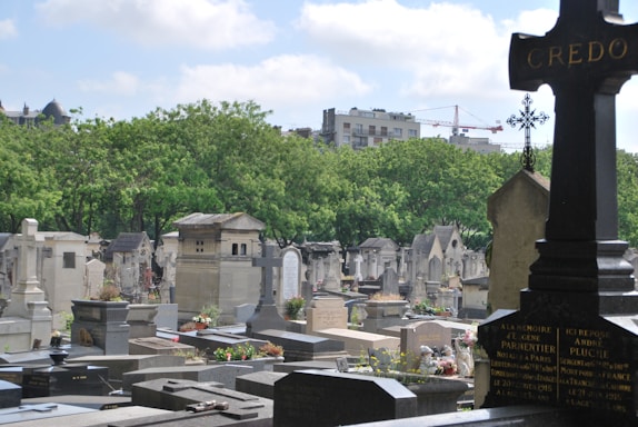A tranquil cemetery scene with numerous tombstones and mausoleums surrounded by lush green trees. In the foreground, there is a prominent black cross with gold lettering. The background reveals a cityscape with buildings and a crane visible under a partly cloudy sky.