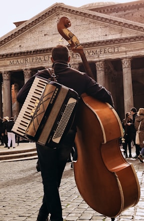 A street musician carries both an accordion and a double bass, standing in front of an ancient building with large columns and the inscription 'MAGRIPPAFECIT'. The scene is bustling with people in the background on a cobblestone path.