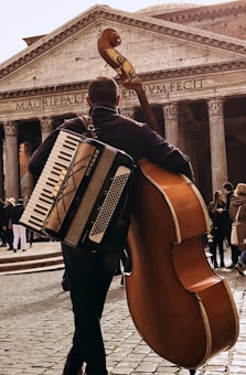 A street musician carries both an accordion and a double bass, standing in front of an ancient building with large columns and the inscription 'MAGRIPPAFECIT'. The scene is bustling with people in the background on a cobblestone path.