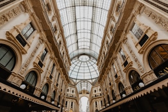 An elegant indoor shopping gallery with a vaulted glass ceiling and ornate architectural details on the surrounding walls. The building features arches, intricate moldings, and large windows, creating a luxurious and historic ambiance.