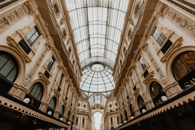 An elegant indoor shopping gallery with a vaulted glass ceiling and ornate architectural details on the surrounding walls. The building features arches, intricate moldings, and large windows, creating a luxurious and historic ambiance.