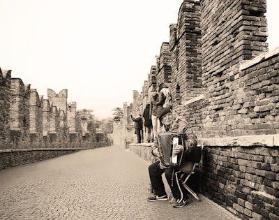 A street musician sitting on a chair, playing an accordion along a historic brick wall. Several people are walking in the background, away from the musician, along a cobblestone path. The scene is monochromatic, with the focus on the musician expressive in his performance.