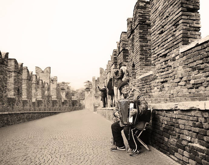 A street musician sitting on a chair, playing an accordion along a historic brick wall. Several people are walking in the background, away from the musician, along a cobblestone path. The scene is monochromatic, with the focus on the musician expressive in his performance.