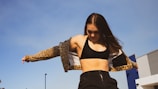 A woman wearing a black sports bra and leopard print jacket is standing outdoors against a clear blue sky. Her long hair is flowing, and she appears to be in motion, adjusting her jacket. The background includes a modern building structure with metal and blue elements.