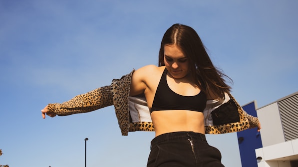 A woman wearing a black sports bra and leopard print jacket is standing outdoors against a clear blue sky. Her long hair is flowing, and she appears to be in motion, adjusting her jacket. The background includes a modern building structure with metal and blue elements.