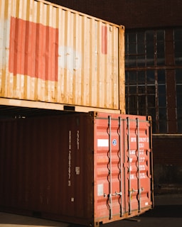 Stacked shipping containers in an industrial area with one red and one beige container. They are positioned in front of a large building with old, multi-paned windows.