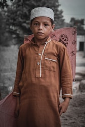 A young boy stands outdoors holding a large kite. He is wearing a brown traditional outfit and a white cap. The background is blurred, suggesting a rural setting with trees and a building.