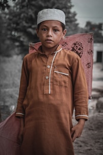A young boy stands outdoors holding a large kite. He is wearing a brown traditional outfit and a white cap. The background is blurred, suggesting a rural setting with trees and a building.