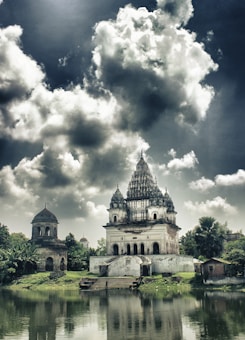 A grand, historic temple structure with multiple domes and ornate architecture sits beside a calm water body. The sky above is dramatic with large, dark clouds creating a moody atmosphere. Lush greenery surrounds the temple, and reflections of the building and the foliage can be seen in the water.