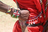 A close-up of an arm adorned with colorful bead bracelets, including one with the Kenyan flag design, rests on a person's back. The individual wears a red garment with decorative beads and metallic discs. The scene suggests cultural attire and craftsmanship.