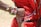 A close-up of an arm adorned with colorful bead bracelets, including one with the Kenyan flag design, rests on a person's back. The individual wears a red garment with decorative beads and metallic discs. The scene suggests cultural attire and craftsmanship.