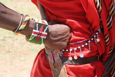 A close-up of an arm adorned with colorful bead bracelets, including one with the Kenyan flag design, rests on a person's back. The individual wears a red garment with decorative beads and metallic discs. The scene suggests cultural attire and craftsmanship.