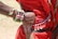 A close-up of an arm adorned with colorful bead bracelets, including one with the Kenyan flag design, rests on a person's back. The individual wears a red garment with decorative beads and metallic discs. The scene suggests cultural attire and craftsmanship.