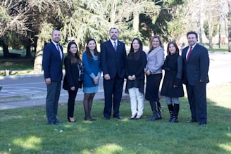 A group of eight individuals is standing on a grassy area outdoors. They are dressed in formal and semi-formal attire, suggesting a professional or official gathering. The individuals appear to be posing for a group photo, standing in a line with some trees and a road visible in the background.