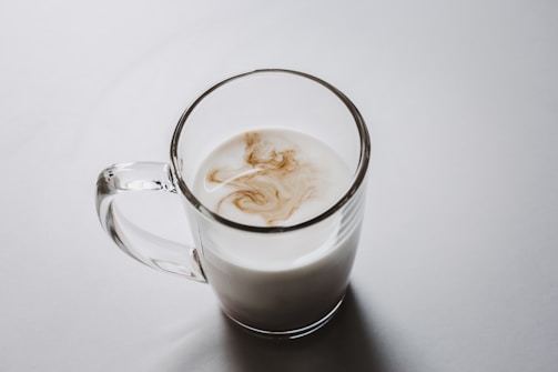 A clear glass mug filled with milk, with a swirl of coffee or tea creating a marbled pattern on the surface.