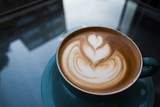 A cup of coffee with intricate latte art featuring a floral design on the frothy surface. The coffee is served in a teal cup and saucer, placed on a reflective glass table. The background shows subtle reflections of buildings and windows.