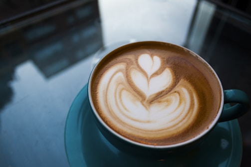 A cup of coffee with intricate latte art featuring a floral design on the frothy surface. The coffee is served in a teal cup and saucer, placed on a reflective glass table. The background shows subtle reflections of buildings and windows.