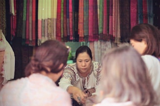 A woman is engaged in a conversation or transaction, surrounded by people, in front of a colorful array of woven textiles hanging in the background. She is dressed in a traditional embroidered blouse, focused on interacting with others, who are partially visible from behind.