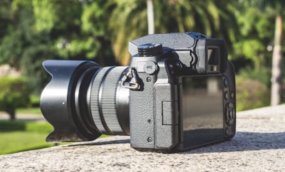 A professional DSLR camera rests on a flat stone surface with a blurred background of lush green trees and grass. The camera is viewed from the back with the LCD screen visible and the lens extending to the left.