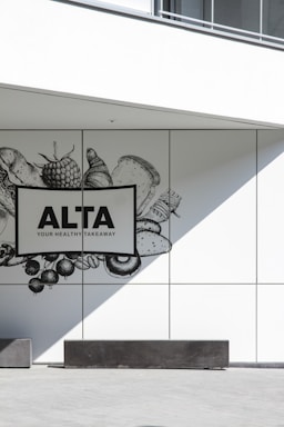 A minimalistic wall design featuring a monochrome illustration of various food items such as vegetables, fruits, and bread. The focus is on a sign with bold text reading 'ALTA YOUR HEALTHY TAKEAWAY'. The scene is complemented by sharp shadows and clean lines, creating a modern and sleek look.