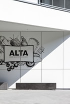 A minimalistic wall design featuring a monochrome illustration of various food items such as vegetables, fruits, and bread. The focus is on a sign with bold text reading 'ALTA YOUR HEALTHY TAKEAWAY'. The scene is complemented by sharp shadows and clean lines, creating a modern and sleek look.