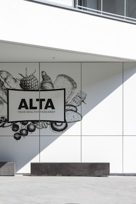 A minimalistic wall design featuring a monochrome illustration of various food items such as vegetables, fruits, and bread. The focus is on a sign with bold text reading 'ALTA YOUR HEALTHY TAKEAWAY'. The scene is complemented by sharp shadows and clean lines, creating a modern and sleek look.