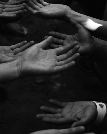 A group of outstretched hands reaching out in different directions. The image is in black and white, creating a dramatic and introspective effect. The hands are of varying skin tones and sizes, suggesting diversity and unity.