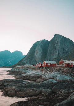 Red cabins stand on stilts along a rocky shoreline, set against towering mountains in the background. The sky is clear, and the water is calm at the edge of the shore.