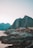 Red cabins stand on stilts along a rocky shoreline, set against towering mountains in the background. The sky is clear, and the water is calm at the edge of the shore.