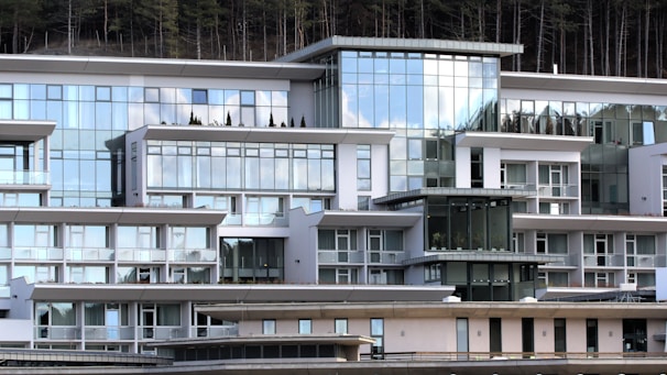 A modern, multi-story building with expansive glass windows and a geometric design. The architectural style features clean lines and a white facade, complemented by balconies and terraces. Reflective surfaces capture the sky and surrounding environment, adding to the contemporary aesthetic. Sparse, neatly arranged shrubbery is visible on some terraces.