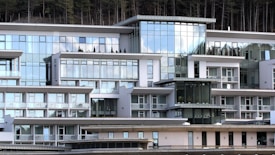 A modern, multi-story building with expansive glass windows and a geometric design. The architectural style features clean lines and a white facade, complemented by balconies and terraces. Reflective surfaces capture the sky and surrounding environment, adding to the contemporary aesthetic. Sparse, neatly arranged shrubbery is visible on some terraces.