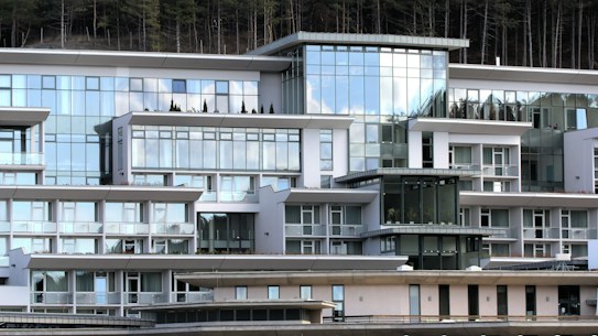 A modern, multi-story building with expansive glass windows and a geometric design. The architectural style features clean lines and a white facade, complemented by balconies and terraces. Reflective surfaces capture the sky and surrounding environment, adding to the contemporary aesthetic. Sparse, neatly arranged shrubbery is visible on some terraces.