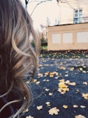 Blonde curly hair is prominently featured in the foreground, while in the background, a paved surface is scattered with fallen autumn leaves, and a building is partially visible.