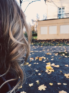 Blonde curly hair is prominently featured in the foreground, while in the background, a paved surface is scattered with fallen autumn leaves, and a building is partially visible.