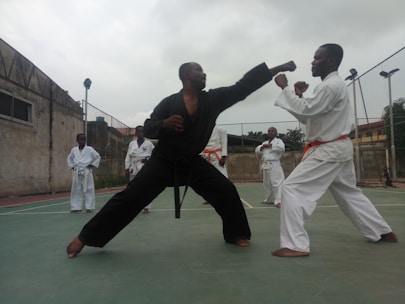 Two individuals are engaged in a martial arts sparring session, wearing traditional uniforms. The person on the left is in a black uniform, executing a punch, while the person on the right, wearing a white uniform with an orange belt, assumes a defensive stance. Other participants in white uniforms are observing in the background on an outdoor court enclosed by a wire fence and adjacent to an old building.