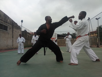 Two individuals are engaged in a martial arts sparring session, wearing traditional uniforms. The person on the left is in a black uniform, executing a punch, while the person on the right, wearing a white uniform with an orange belt, assumes a defensive stance. Other participants in white uniforms are observing in the background on an outdoor court enclosed by a wire fence and adjacent to an old building.