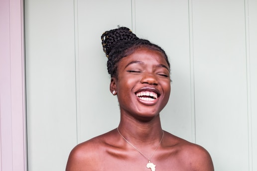 A person with braided hair is laughing joyfully. They are wearing a simple necklace with a pendant shaped like a continent. The background is a light green vertical panel.