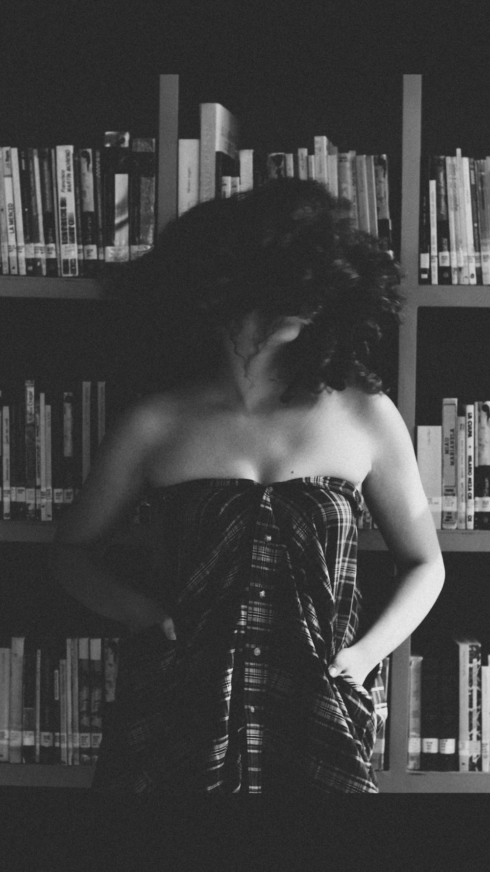 fotografia in scala di grigi di donna che indossa pantaloni marroni con capelli ricci in piedi accanto alla libreria