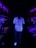 A person stands in a dimly lit hallway illuminated by ultraviolet light, wearing a floral patterned shirt and white shoes that glow brightly. On both sides, aquarium displays with coral and sea plants are visible, producing a vibrant neon effect with purples, reds, and greens.
