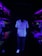 A person stands in a dimly lit hallway illuminated by ultraviolet light, wearing a floral patterned shirt and white shoes that glow brightly. On both sides, aquarium displays with coral and sea plants are visible, producing a vibrant neon effect with purples, reds, and greens.