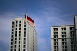 Tall white building with many windows stands beside a flagpole. The red and white flag is waving against a backdrop of blue sky with scattered clouds.