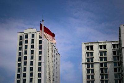 Tall white building with many windows stands beside a flagpole. The red and white flag is waving against a backdrop of blue sky with scattered clouds.