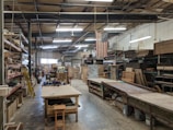An industrial workspace with high ceilings and fluorescent lighting. Wooden shelves with various materials and tools line the walls. There are long workbenches in the middle of the room, and an American flag hangs prominently. The area appears organized with a large number of wooden and metal objects, along with stacked wooden boards.
