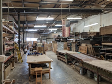 An industrial workspace with high ceilings and fluorescent lighting. Wooden shelves with various materials and tools line the walls. There are long workbenches in the middle of the room, and an American flag hangs prominently. The area appears organized with a large number of wooden and metal objects, along with stacked wooden boards.