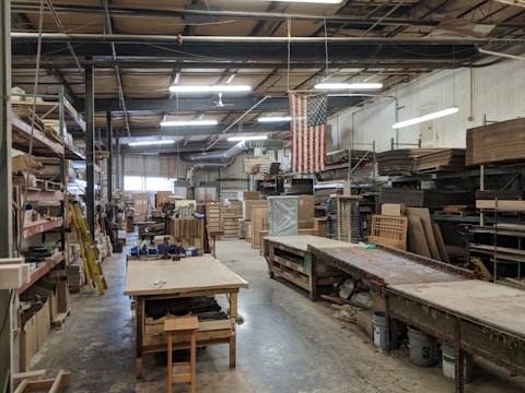An industrial workspace with high ceilings and fluorescent lighting. Wooden shelves with various materials and tools line the walls. There are long workbenches in the middle of the room, and an American flag hangs prominently. The area appears organized with a large number of wooden and metal objects, along with stacked wooden boards.