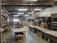 An industrial workspace with high ceilings and fluorescent lighting. Wooden shelves with various materials and tools line the walls. There are long workbenches in the middle of the room, and an American flag hangs prominently. The area appears organized with a large number of wooden and metal objects, along with stacked wooden boards.