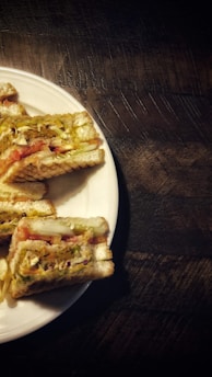 Sandwiches are arranged on a round white plate placed on a dark wooden surface. The sandwiches appear toasted with visible layers of tomato, lettuce, and possibly cheese or other fillings.