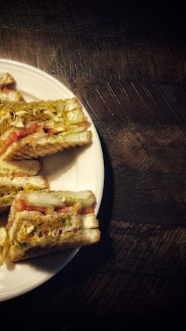 Sandwiches are arranged on a round white plate placed on a dark wooden surface. The sandwiches appear toasted with visible layers of tomato, lettuce, and possibly cheese or other fillings.