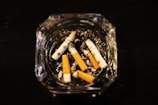 Several cigarette butts are scattered in a glass ashtray filled with ash, positioned on a dark surface.
