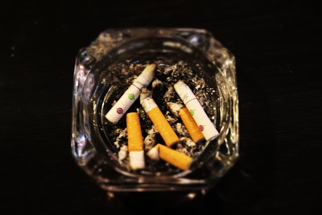 Several cigarette butts are scattered in a glass ashtray filled with ash, positioned on a dark surface.